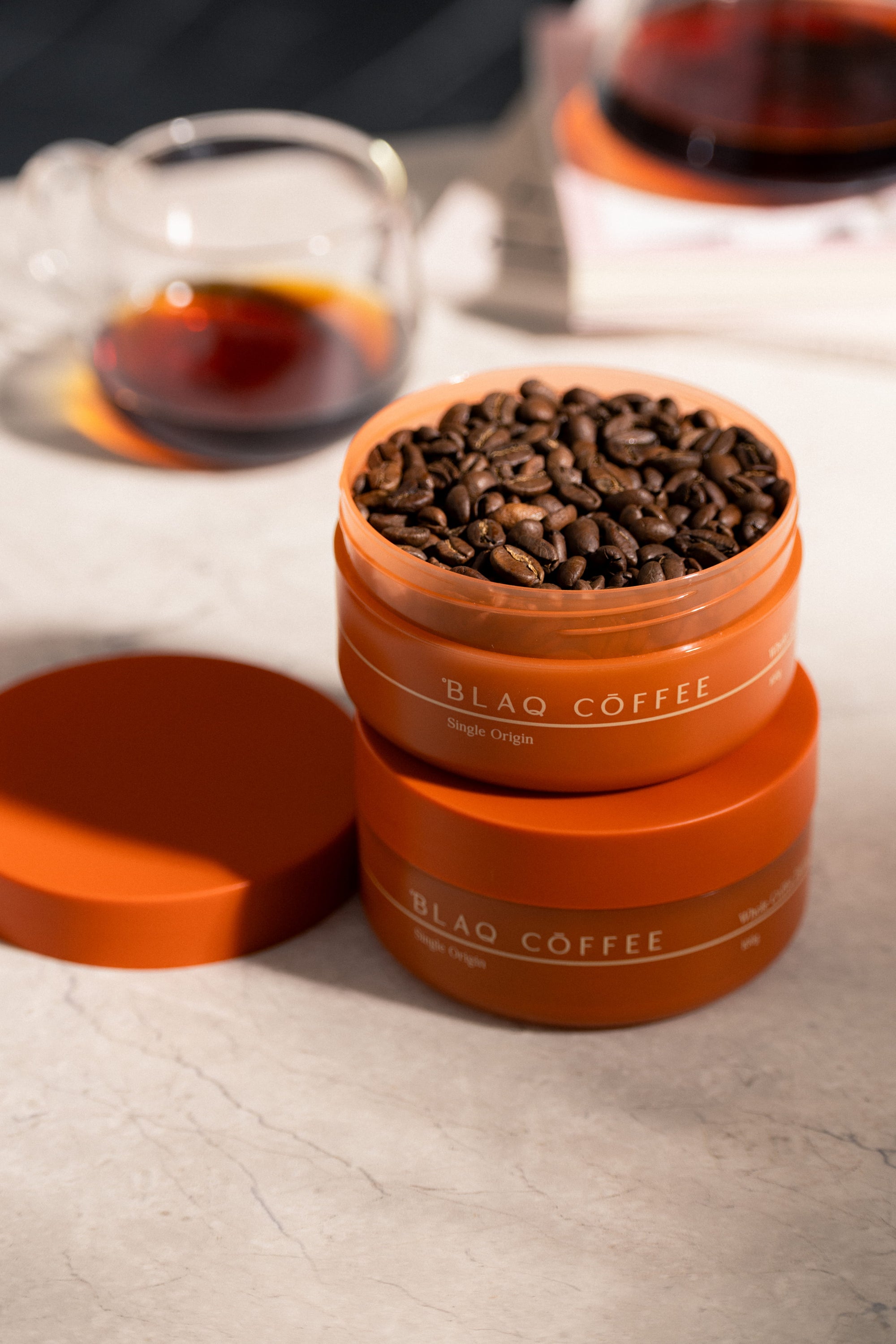 A just-opened Blaq Coffee jar of freshly roasted beans stacked on a second jar, a brewed cup behind.