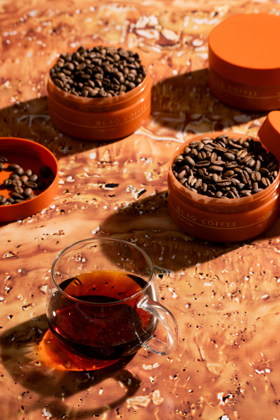 Two open Blaq Coffee jars of roasted beans side by side with a brewed cup, on a warm marbled surface.